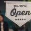 Sign In A Window That States, “Yes, We’re Open” With Customers Blurred In The Background.
